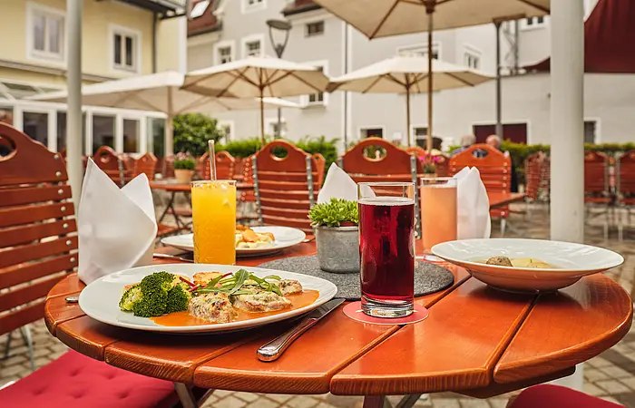 Die Terrasse ist mit Holzmöbeln bestückt. Auf dem vordersten Tisch sind zwei Essen und zwei Getränke angerichtet. Im Hintergrund sieht man die weißen Sonnenschirme.