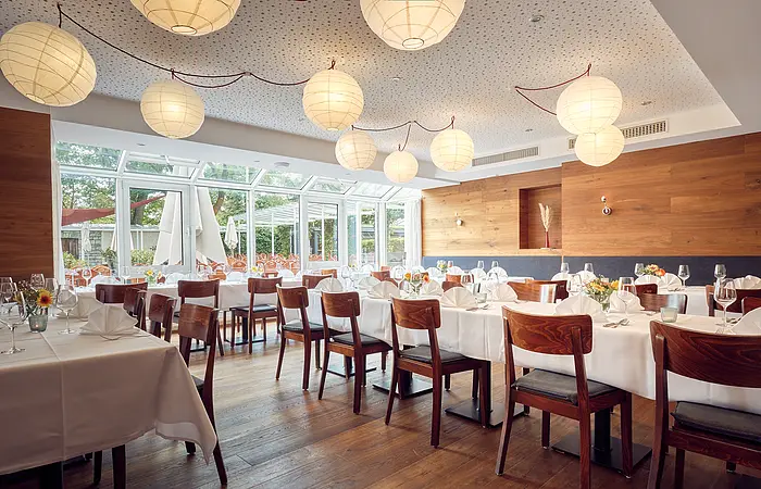 Das Restaurant mit runden, herabhängenden Lampen ist hell und modern eingerichtet. Der dunkle Holzboden passt perfekt zu den Tischen, die mit weißen Tischdecken gedeckt werden. Alle Tische sind festlich gedeckt.