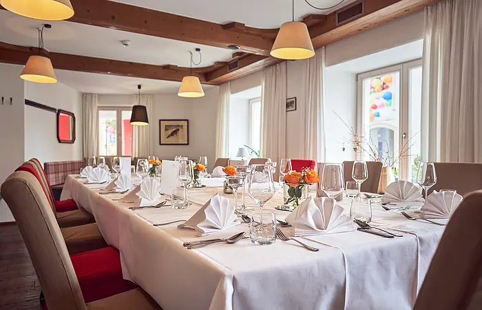 Der Raum mit den herabhängenden Leuchten ist mit einer großen Tafel und braunroten bequemen Stühlen gefüllt. Der Tisch ist festlich mit Tischdecke, weißen Servietten, Geschirr und Weingläsern gedeckt.