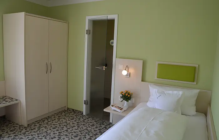 Hotelzimmer mit Einzelbett, grün gestrichener Wand, Schrank, Glastür und Nachttisch mit Lampe und Blumen.