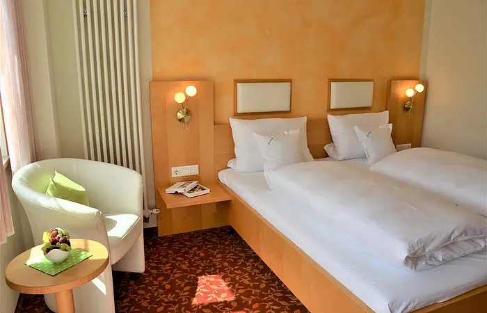 Hotelzimmer mit zwei Einzelbetten, Nachttisch, Sessel, kleinem Tisch und Obstschale bei Tageslicht.