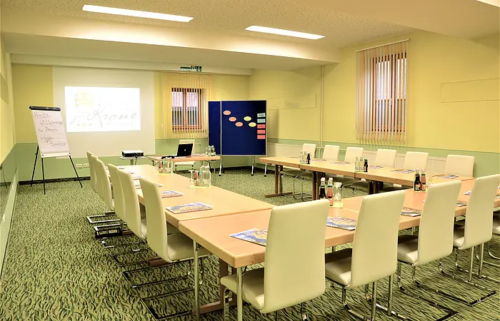 Tagungsraum mit U-förmiger Tischanordnung, weißen Stühlen, Flipchart und Projektor mit Leinwand.