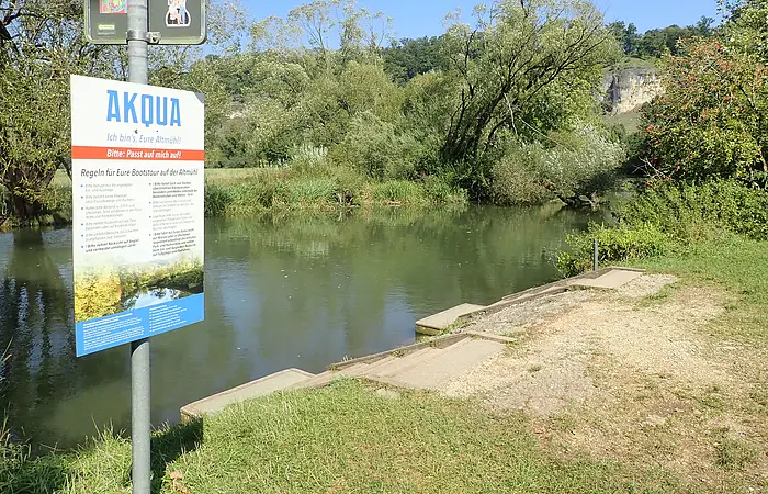 Schild mit Bootstour-Regeln an Flussufer mit Treppe und Bäumen unter blauem Himmel.