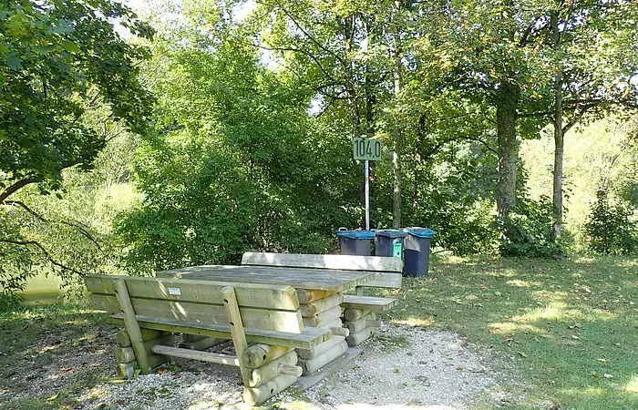 Holztisch mit Bänken im Wald, dahinter drei Mülltonnen und ein Schild.