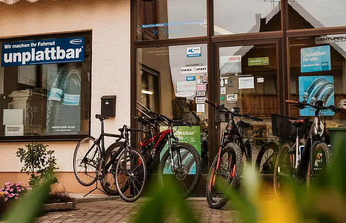 Fahrradgeschäft mit mehreren Fahrrädern vor dem Eingang und Werbeplakaten zu pannensicheren Reifen auf der Glasscheibe des Geschäfts. Im Vordergrund unscharf grüne Pflanzen zu sehen.