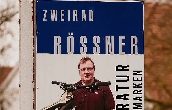 Werbeschild für Fahrradreparatur mit Mann und Fahrrad, Text „Zweirad Rössner Reparatur aller Marken“ und Telefonnummer.