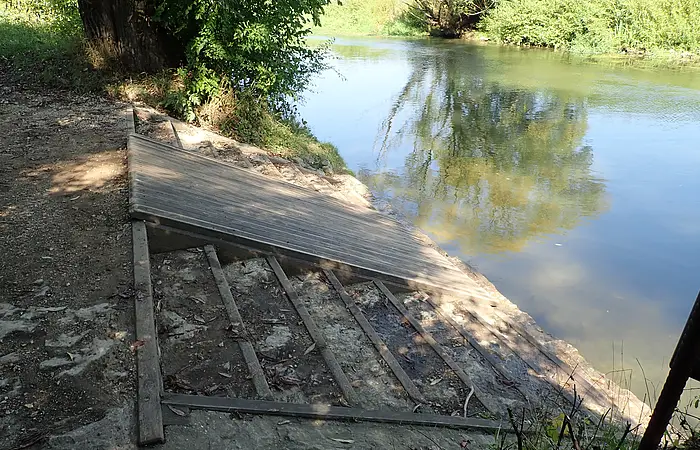Holzrampe mit Stufen am Ufer eines ruhigen Flusses, umgeben von Bäumen und Pflanzen