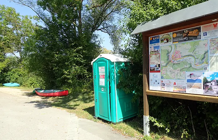 Mobiles Toilettenhäuschen neben Infotafel mit Landkarte und Kanus am Weg in bewaldeter Umgebung.