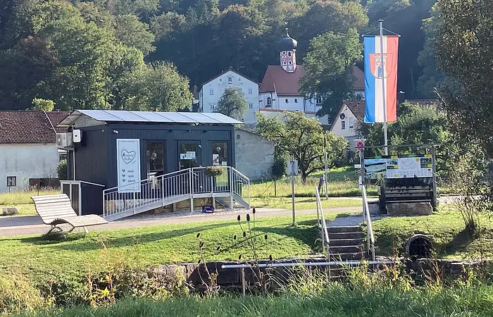 Kleines Gebäude mit Rampe, Bank, Fahne und Kirche im Hintergrund vor bewaldetem Hügel bei Sonnenschein.