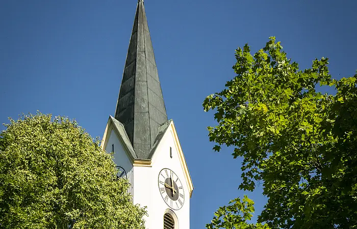 Kirchturm mit Uhr und Kreuz, umgeben von grünen Bäumen, vor blauem Himmel.