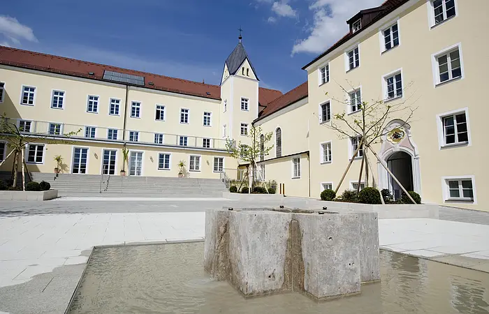 Innenhof mit modernem Brunnen und gelben Gebäuden mit roten Dächern unter blauem Himmel