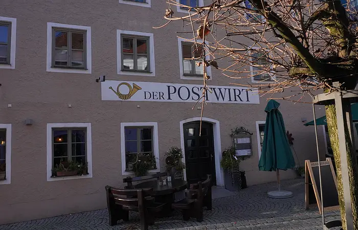 Außenansicht eines Gebäudes mit Schild „Der Postwirt“, Tischen, Stühlen und kahlem Baum im Vordergrund.