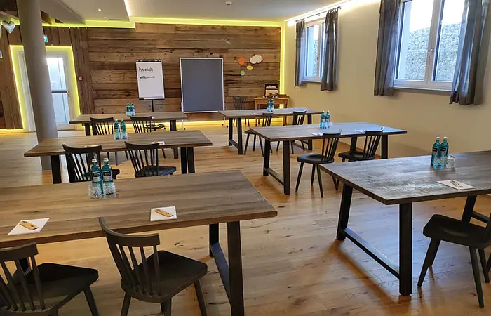 Konferenzraum mit Holztischen, Stühlen, Wasserflaschen und Flipchart vor Holzpaneelwand und Fenstern mit Vorhängen.