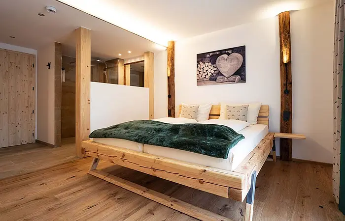 Schlafzimmer mit Holzbett, grünem Überwurf, Holzboden und offener Dusche hinter Holztrennwand.