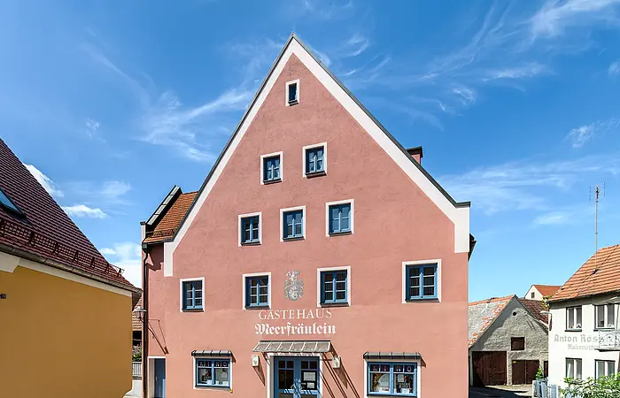 Rosa Gästehaus mit blau gerahmten Fenstern und Eingang unter blauem Himmel in einer Gasse mit Kopfsteinpflaster.