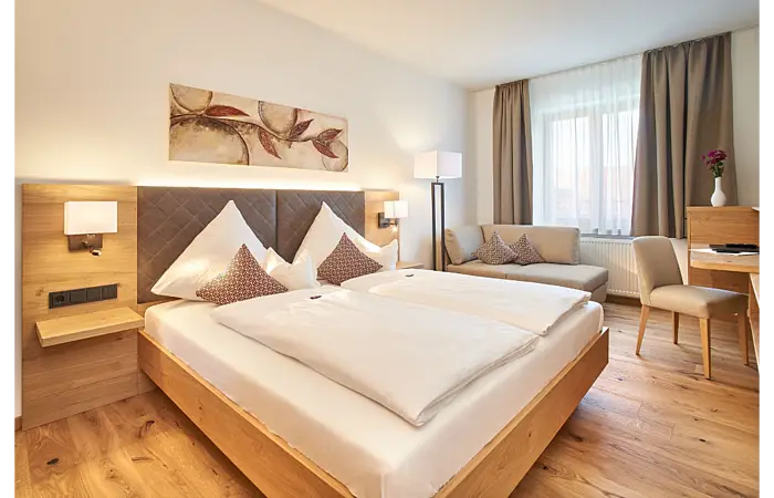 Ein großes Doppelbett steht in einem hellen Raum mit weißen Wänden und braunen Elementen. Über dem Bett hängt ein Bild.