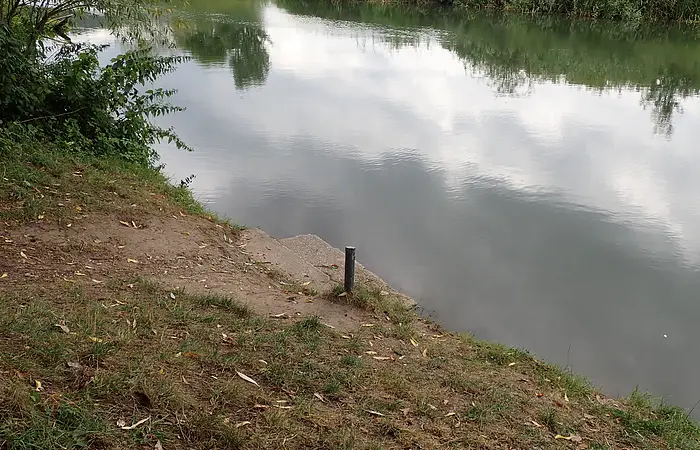 Ufer mit Gras und Erde, kleine Treppe und Pfahl am ruhigen Fluss mit Spiegelung des Himmels und Büschen.