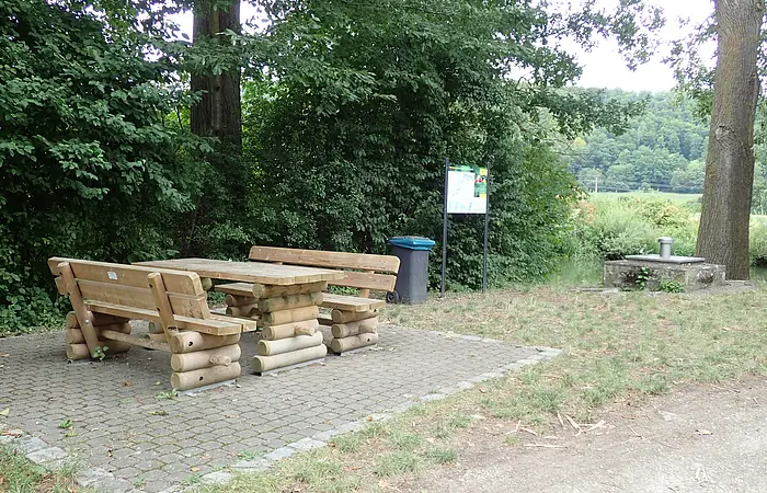 Holztisch mit zwei Bänken auf gepflastertem Platz neben Wald und Infotafel mit Mülleimer.