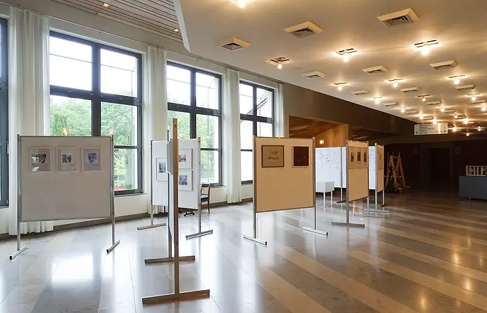 Großer Ausstellungsraum mit mehreren Stellwänden und Kunstwerken vor großen Fenstern und heller Deckenbeleuchtung.