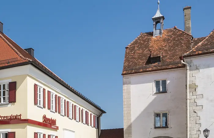 Tourist-Information der Stadt Monheim/Monheimer Alb: ein Fachwerkhaus mit drei Stockwerken, weißen Wänden und braunen Balken. Davor steht ein Schaukasten und zwei Radfahrer fahren auf gepflasterter Straße vorbei. Rechts davon ein gelbes zweistöckiges Gebäude mit roten Fensterläden.