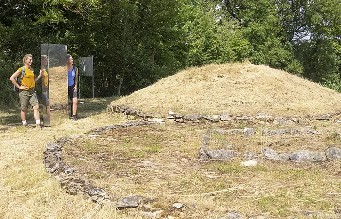 An der Grabhügelanlage stehen zwei Wanderer in sommerlicher Kleidung an einer Stele und bestaunen die Hügelanlage. Hinter ihnen dichter dunkelgrüner Wald.
