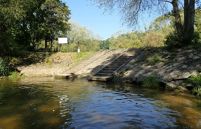 Ufer mit zwei Holzstegen und Treppen, umgeben von Bäumen und Wasser unter blauem Himmel.