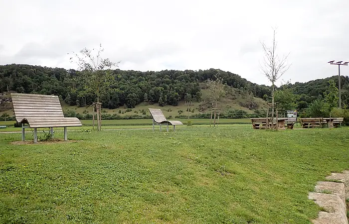 Grüne Wiese mit mehreren Holzliegen und Bänken, im Hintergrund bewaldete Hügel unter bewölktem Himmel.
