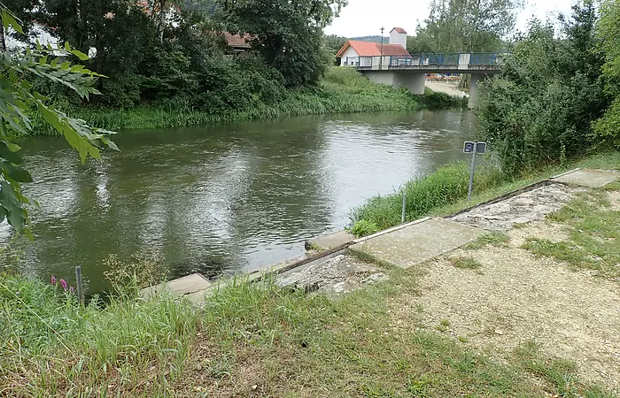 Flussufer mit Büschen und Bäumen, im Hintergrund eine Brücke und ein Haus mit rotem Dach.