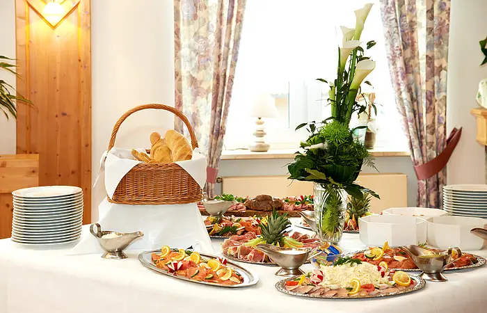 Es leckeres kaltes Buffet ist vor einem Fenster aufgebaut. Es gibt Platten mit Wurst, Lachs und Käse. Erhöht steht der große gut gefüllte Brotkorb. Eine Blumendeko und Teller gehören ebenfalls dazu.