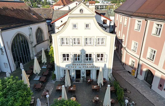 Historisches Gebäude mit Café und Restaurant, umgeben von Tischen und Sonnenschirmen auf einem gepflasterten Platz.