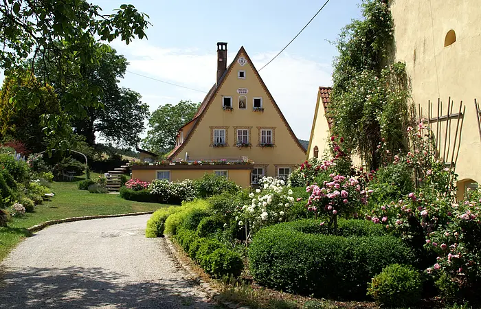 Gepflasterter Weg führt zu gelbem Haus mit Giebeldach, umgeben von grünen Büschen und blühenden Rosen.