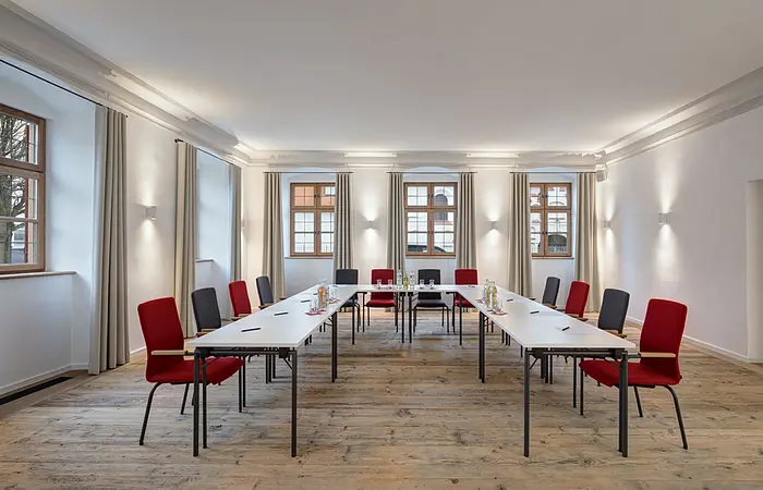 Konferenzraum mit U-förmiger Tischanordnung, roten und grauen Stühlen und Fenstern mit Vorhängen.