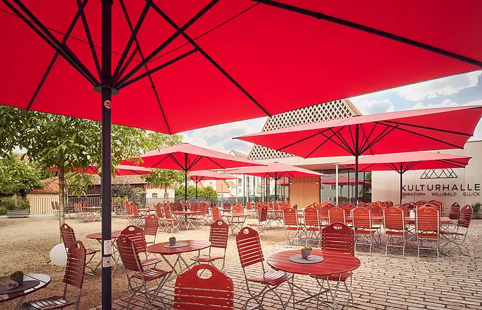 Außenbereich eines Cafés mit roten Sonnenschirmen und roten Stühlen vor einem Gebäude mit Schriftzug Kulturhalle.