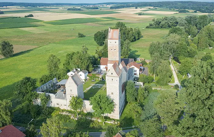 Luftaufnahme einer mittelalterlichen Burg mit drei Türmen umgeben von Feldern und Bäumen an einem sonnigen Tag.