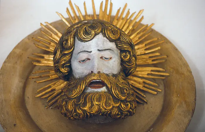 Holzskulptur eines bärtigen Männerkopfes mit geschlossenen Augen, umgeben von goldenen Strahlen.
