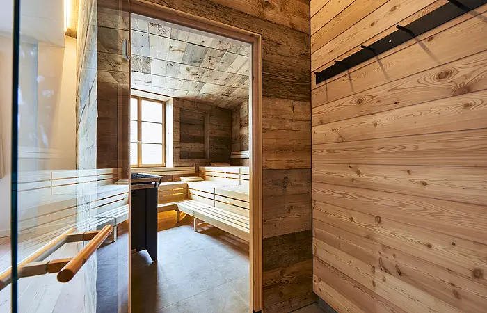 Die Glastür der Sauna aus Holz steht offen und wir können einen Blick auf die leeren Bänke darin werfen. Ein Fenster gegenüber lässt Licht herein.