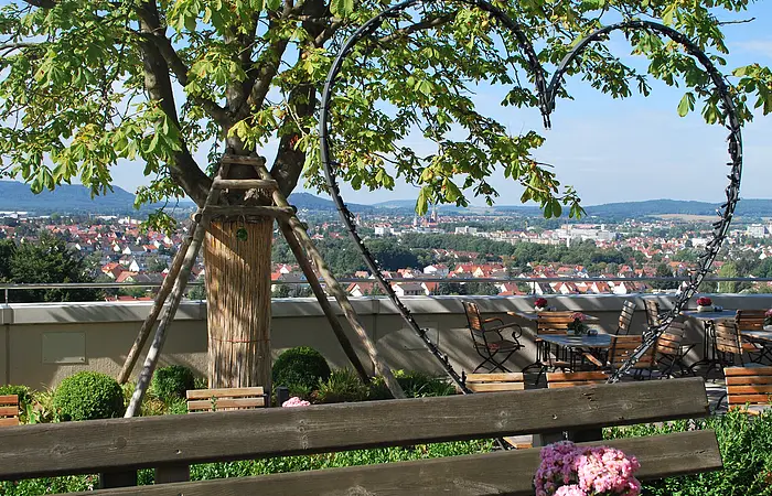 Blick von Terrasse mit Holzbänken und Tischen auf Stadt, Baum mit Stütze und Herz aus Metall im Vordergrund.