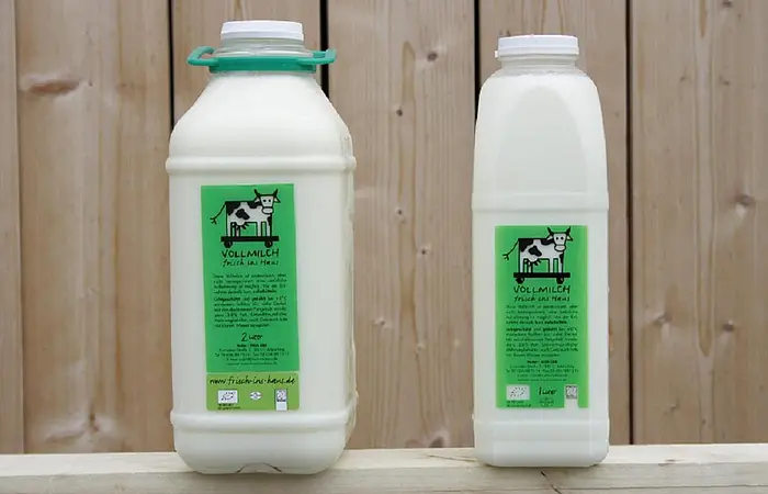 Zwei weiße Plastikmilchflaschen mit grünem Produktaufkleber stehen auf einem weißen Brett. Die Milchflaschen haben verschiedene Größen, 1l und 0,5 l. Im Hintergrund sieht man eine Holzwand.