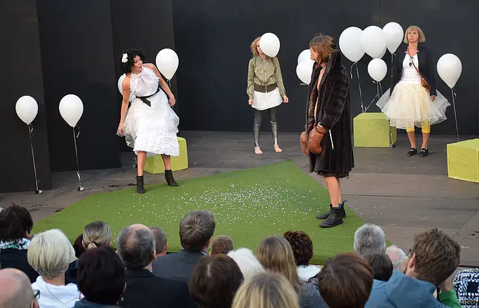 Zuschauer sitzen vor einer Bühne. Auf der Bühne wird ein Theaterstück aufgeführt. Die Bühne ist geschmückt mit weißen Ballons. Die vier Schauspieler auf der Bühne tragen weiße Tüllkleider.