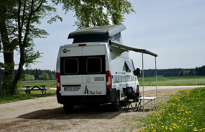 Wohnmobil mit ausgefahrener Markise steht auf einem Feldweg neben einer Wiese mit Löwenzahn.