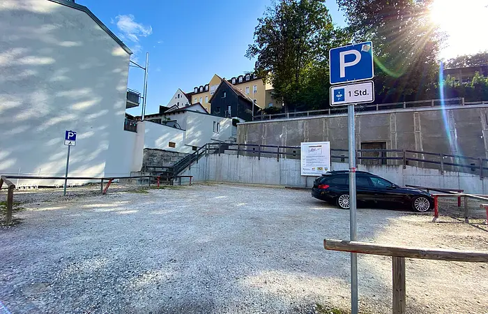 Parkplatz mit Schotterboden, zwei Parkschildern und einem schwarzen Auto, umgeben von Gebäuden und Bäumen