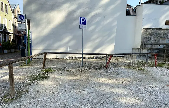 Parkplatz für Menschen mit Behinderung vor weißer Hauswand mit Absperrung aus Holzpfosten und Metallstange