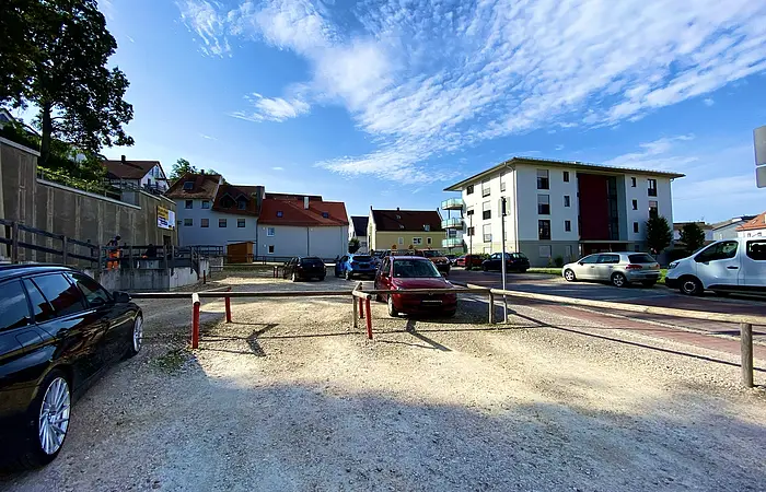 Parkplatz mit mehreren geparkten Autos vor Wohnhäusern unter blauem Himmel mit Wolken