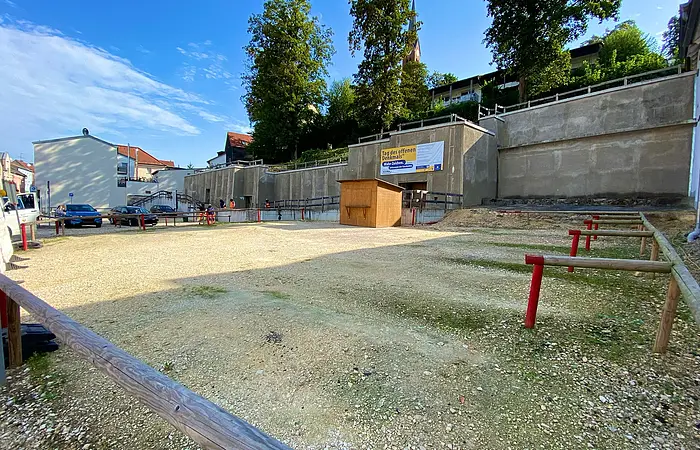 Freier, eingezäunter Platz mit Kiesboden vor einer Betonmauer und einem Holzhäuschen unter blauem Himmel.