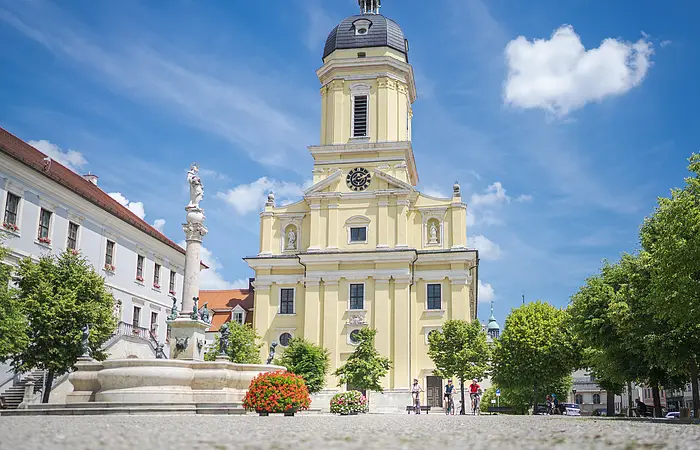 Radelnde auf dem Karlsplatz vor der prächtigen Hofkirche. Im Vordergrund ist ein Brunnen zu sehen.