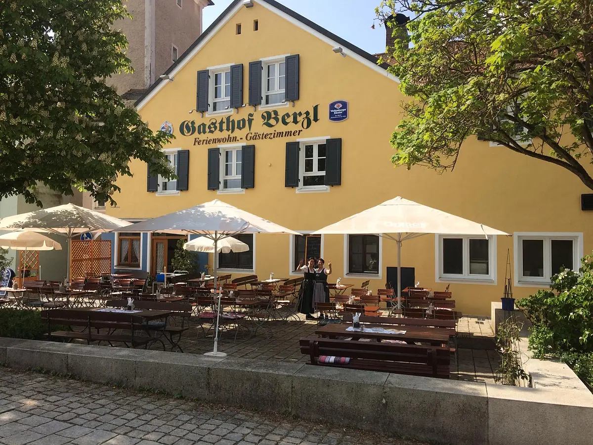 Gasthof Berzl Außenbereich mit Tischen, Stühlen und Sonnenschirmen vor gelbem Gasthof mit Schriftzug „Gasthof Berzl“.