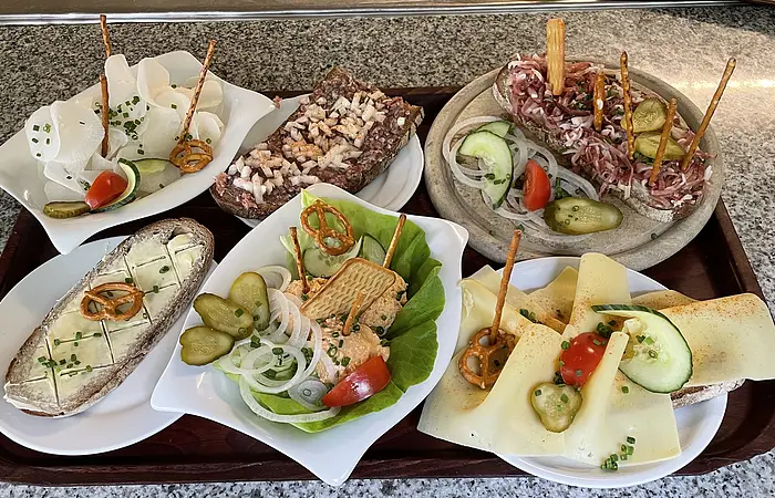 Auf einem Tablett sind verschiedene Brotzeiten wie Butterbrot, Wurstsalat, Käsebrot, Schinkenbrot, Rettich und Tartarbrot zu sehen.