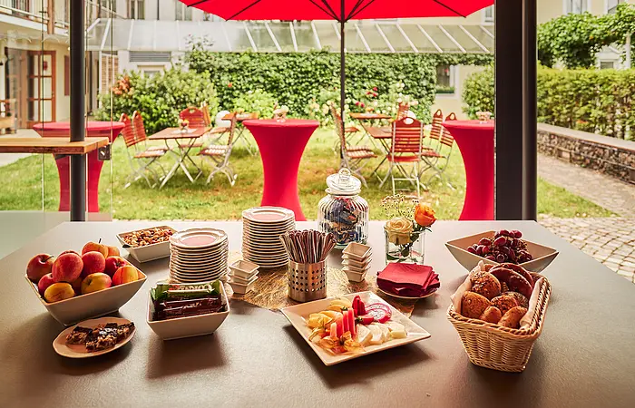 Buffet mit Obst, Gebäck, Nüssen und Tellern auf Tisch vor Garten mit roten Stehtischen und Stühlen