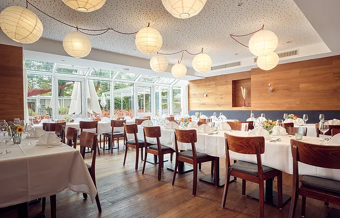 Restaurant mit gedeckten Tischen, Holzstühlen, großen Fenstern und runden Papierlampen an der Decke.