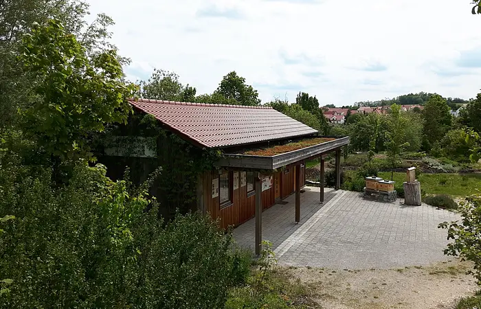 Holzgebäude mit rotem Dach und begrüntem Vordach, umgeben von Bäumen und einem gepflasterten Vorplatz, bei bewölktem Himmel.